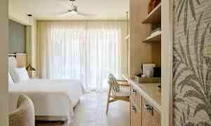 Et hotelværelse med en stor seng, en lænestol og hylder med kaffemaskine, oplyst af naturligt lys gennem store gardiner. (AI-genereret tekst)