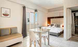 Et lyst hotelværelse med en seng, et spisebord med to stole og en lille sofa. Vindue med gardin og badeværelse ses i baggrunden. (AI-genereret tekst)