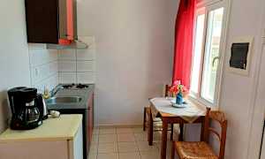 Et lille køkken med en håndvask og komfur samt et bord med stole ved et vindue. (AI-genereret tekst)