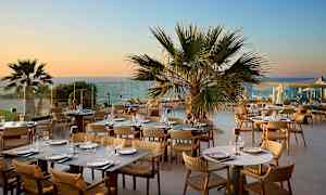 En udendørs restaurant med borde og stole, omgivet af palmer og med udsigt til havet i solnedgangen. (AI-genereret tekst)