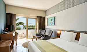 Et hotelværelse med en dobbeltseng, to stole, et bord og et TV-møbel. En balkon med udsigt til palmer er synlig gennem en glasdør. (AI-genereret tekst)