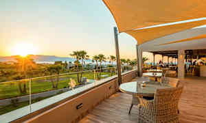 Terrasse med borde og stole samt udsigt over grønne områder og solnedgang. (AI-genereret tekst)