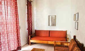 Stuen har røde gardiner, en træsofa med røde ternede puder og en stofbetrukket stol ved siden af. (AI-genereret tekst)