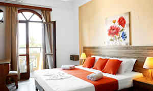 Et hotelværelse med en seng dækket med hvide og orange lagner, et træ-skrivebord og en åbning mod en balkon. (AI-genereret tekst)