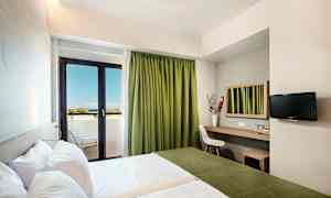 Et hotelværelse med to senge, en arbejdsplads med spejl, en stol og et tv på væggen. Værelset har en balkon med grønne gardiner og udsigt til havet. (AI-genereret tekst)