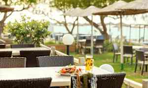 Et bord med en tallerken salat og et glas øl på en terrasse med havemøbler og strandparasoller i baggrunden. (AI-genereret tekst)