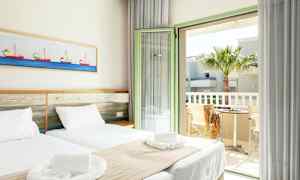 Et lyst hotelværelse med to enkeltsenge, hvidt sengetøj og beige sengetæpper. Et billede med bådmotiver hænger på væggen over sengen. Værelset har en åben altandør med udsigt til en palme og et bord med to stole. (AI-genereret tekst)
