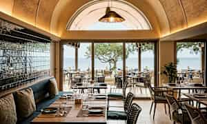 Restaurant med udsigt over havet, møbler i træ og glasdetaljer. (AI-genereret tekst)