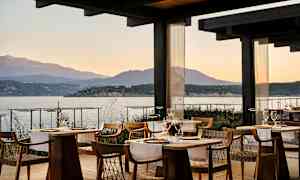 En restaurant med træborde og stole, beliggende ved havet med udsigt over bjerge i baggrunden under solnedgangen. (AI-genereret tekst)