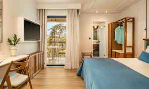 Hotelværelse med dobbeltseng, skrivebord og TV. Glasdøre fører ud til en balkon. Badeværelse synligt gennem en åben dør. (AI-genereret tekst)