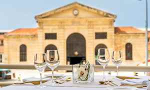 Et dækket bord udendørs med vinglas, bestik og servietter. I baggrunden ses en historisk stenbygning. (AI-genereret tekst)