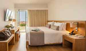 Et hotelværelse med en stor dobbeltseng, skrivebord og åbne døre til en balkon med udsigt. (AI-genereret tekst)