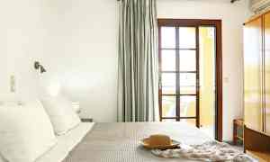 Et soveværelse med en seng, puder og ét tæppe. Et vindue med gardiner og et natbord er synligt. (AI-genereret tekst)