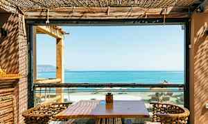 En udendørs terrasse med bord og stole, udsigt over havet og stranden gennem en ramme. (AI-genereret tekst)