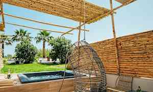 Billede af en terrasse med spabad, hængestol og havemøbler, omgivet af træer og buske. (AI-genereret tekst)