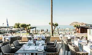 Udendørs servering med borde og stole, havudsigt og strand i baggrunden. (AI-genereret tekst)