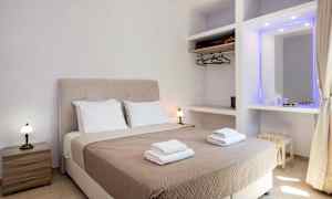 Et hotelværelse med en dobbeltseng udstyret med beige sengetæppe og to sæt håndklæder, samt sidebord og garderobe. (AI-genereret tekst)