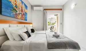 Dobbeltværelse med hvide vægge, en dobbeltseng med gråt sengetæppe og et maleri af sejlbåde på væggen. (AI-genereret tekst)