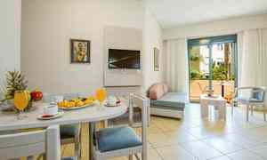 Et lyst hotelværelse med spisebord og bagværk på en tallerken, sofagruppe og en altan i baggrunden. (AI-genereret tekst)