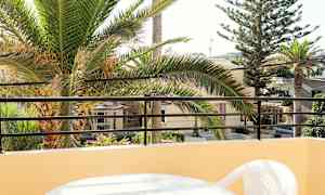 Udsigt fra altan med et bord og plastikstol, palmetræer og bygninger i baggrunden. (AI-genereret tekst)