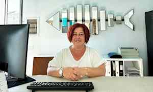 Person sidder ved et skrivebord med tastatur, computerskærm og hylder i baggrunden. (AI-genereret tekst)