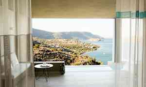 Udsigten fra et værelse med gardiner mod et bjergrigt landskab ved kysten og havet. (AI-genereret tekst)