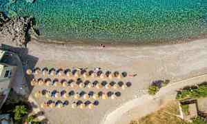 Luftfoto af en strand med opstillede liggestole og parasoller ved kanten af turkisblåt vand. (AI-genereret tekst)