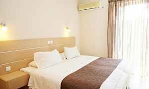 Et hotelværelse med en stor seng, hvidt sengetøj og brunt sengetæppe. Et klimaanlæg er monteret på væggen ved siden af vinduet med lyse gardiner. (AI-genereret tekst)