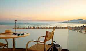 Udsigt fra en terrasse mod havet med solnedgang og strandparasoller i baggrunden. (AI-genereret tekst)