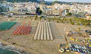 En strand med parasoller og liggestole opstillet i rækker foran en by med mange bygninger. (AI-genereret tekst)