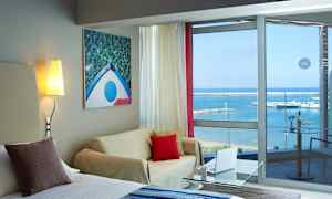 Et hotelværelse med siddeområde, lysmaleri på væggen og glasvinduer med udsigt over havet og en lystbådehavn. (AI-genereret tekst)