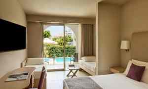 Et hotelværelse med en dobbeltseng, et skriveplads og en ekstra seng. Udenfor ses en terrasse og en pool. (AI-genereret tekst)