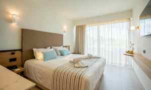 Et hotelværelse med en dobbeltseng, pyntet med puder og rullede håndklæder, vindue med gardiner samt en vægmonteret hylde med en flaske og frugt. (AI-genereret tekst)