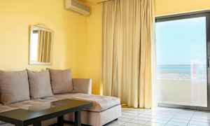 En stue med beige hjørnesofa, glasbord, spejl på væggen og stor vinduesdør med gardiner og udsigt. (AI-genereret tekst)