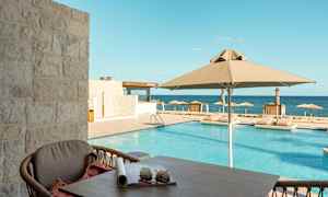 Udsigt fra en terrasse mod en pool med parasoller og liggestole, med havet i baggrunden. (AI-genereret tekst)