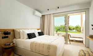 Interiør af et lyst hotelværelse med en dobbeltseng og en lille siddegruppe udenfor med udsigt til grønt. (AI-genereret tekst)