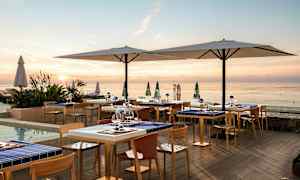 En udendørsrestaurant ved havet ved solnedgang med borde og stole samt store parasoller. (AI-genereret tekst)