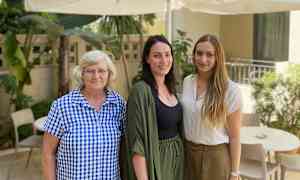 Tre personer står udenfor ved et bord og stole under en parasol. (AI-genereret tekst)