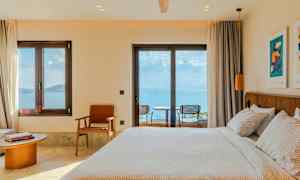 Et hotelværelse med en opredt seng, stole og et bord ved vinduet med udsigt over havet. (AI-genereret tekst)