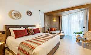 Et hotelværelse med en dobbeltseng, dekoreret med røde og beige tekstiler, og et siddeområde ved vinduet med naturligt lys. (AI-genereret tekst)