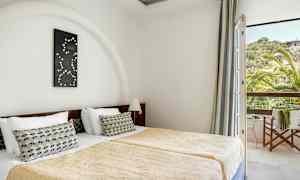 Værelset viser to enkeltsenge med guldfarvede sengetæpper og en dekorativ pude på hver seng. En dekoration hænger over hovedgærdet, og et vindue med udsigt til grønt og et hus ses til højre. (AI-genereret tekst)