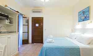 Et hotelværelse med to senge beklædt med blå sengelinned, en træbeklædt dør, et lille køkken med vask og et maleri på væggen. (AI-genereret tekst)