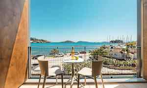 Udsigt fra en balkon med to stole og et bord, mod havet og en lystbådehavn. (AI-genereret tekst)