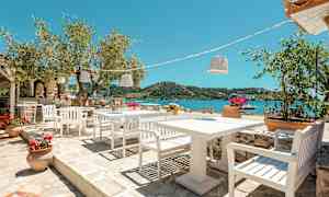 Udendørs servering med hvide stole og borde, udsigt over havet og grønne bakker. (AI-genereret tekst)