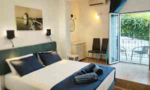 Et hotelværelse med en opredt seng og mørkeblå sengetæpper, et åbent vindue mod altanen, samt møbler inklusive to stole og en kommode. (AI-genereret tekst)