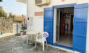 En hvid bygning med blå døre og skodder. Foran døren står to hvide plastikstole og et lille bord. (AI-genereret tekst)