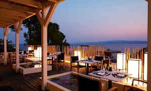 En restaurantterrasse med borde og stole ved solnedgang, havet i baggrunden. (AI-genereret tekst)