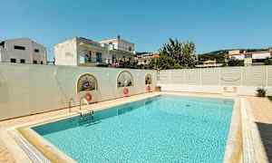 En rektangulær swimmingpool omgivet af en lys terrasse og en lav hvid mur, med bygninger og træer i baggrunden. (AI-genereret tekst)