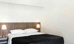 Et soveværelse med en dobbeltseng, hvidt sengetøj og et mørkt sengetæppe. To sengelamper står på sengeborde ved siden af sengen. (AI-genereret tekst)