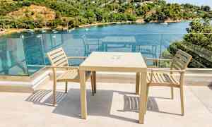 To beige stole og et bord på en terrasse med glasrækværk og udsigt over en bugt med både og grøntområde. (AI-genereret tekst)